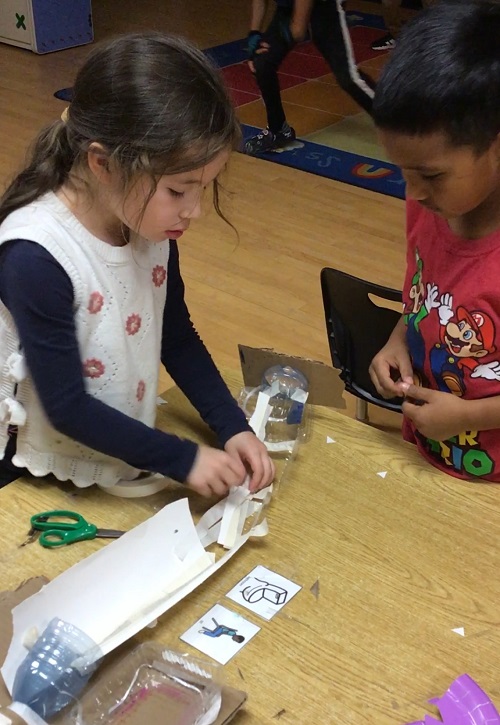 two children at a table building a marble run out of cardboard, tape and recyclables
