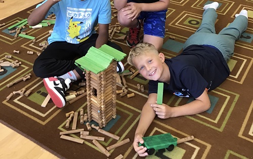 boy laying on the floor looking up smiling, beside a stucture he built with small building logs