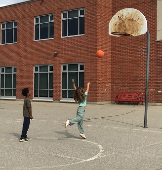 2 children, one shooting a basketball into the basketball net one behind her watching