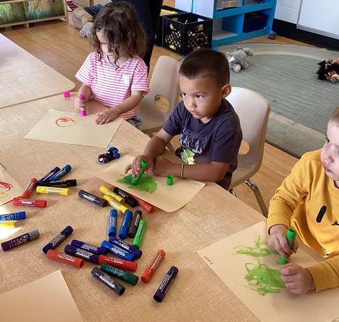 3 children sitting at a table using paint sticks to colour with