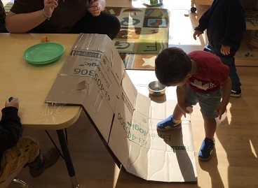 child bending down with a ball in thier hand standing in from on a ramp made out of cardboard, attached to a table