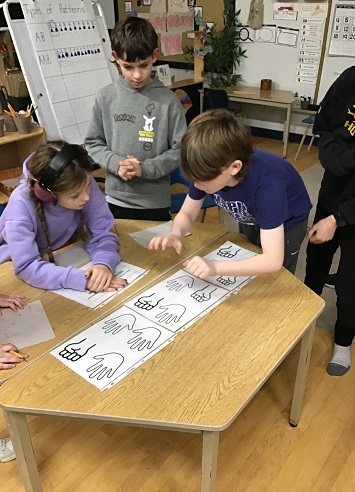 4 School Age children standing by a table with different hand positions, one school age is attempting to match their hands with the paper on the table