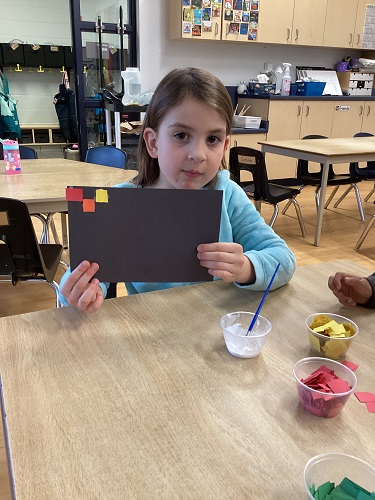 school age child sitting at the table with a black piece of paper and 3 coloured squares glued onto the corner of the paper looking at the camera
