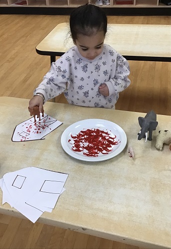 toddler child standing at the table with a toy animal in her hand and a plate of paint and putting the animal on the paper to create footprints