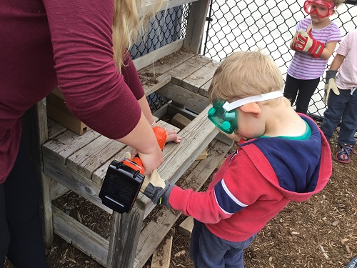 Preschool educatorr and chilld  working together using a drill to fix a wooden mud kitchen