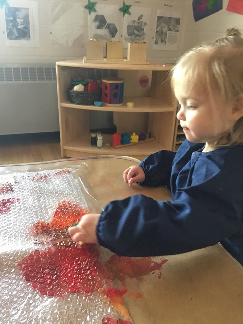 Toddler girl using her finger to paint on bubble wrap using the colours red, brown and orange