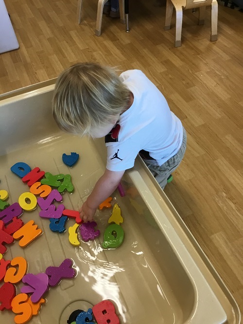 Toddler boy exploring sensory play in a bin filled with foam letters and water
