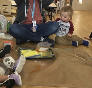 Toddler boy sitting beside an educator observing the science experiment she is conducting  in a ziploc bag that is mixing vinegar and baking soda