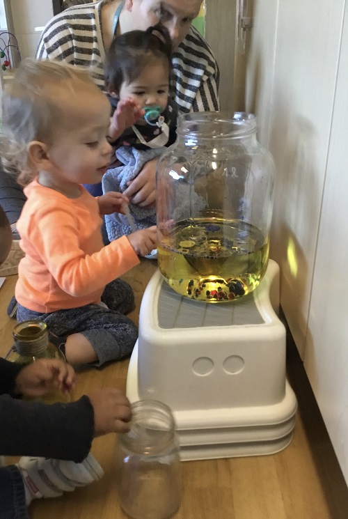 Two infant children sitting with an educator looking into a large pickle jar filled with oil and coloured bubbles