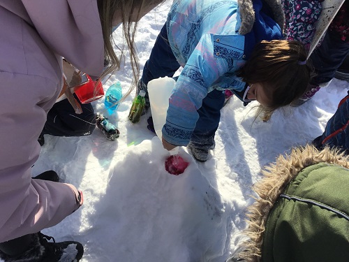 School Age girl adds viengar to the red food colouring in the hole at the top of the snow mound