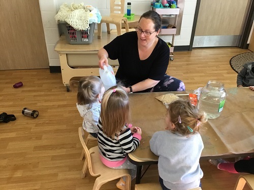 Educator sitting with a small group of toddler children pouring vinegar into a large pickle jar for a science experiment with help from a toddler child