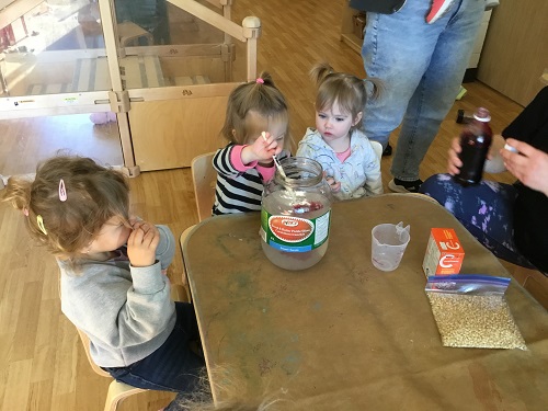 Toddler child sitting at a table with peers stirring a food colouring and vinegar in a large pickle jar 