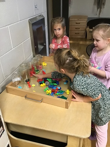 Three children looking at the colourful loose parts on a tray and sorting them into jars