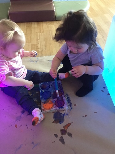 Two infants sitting on the floor with paint and paint brushes in front of them