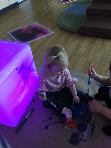 An infant sitting beside a light cube with a paint brush in their hand