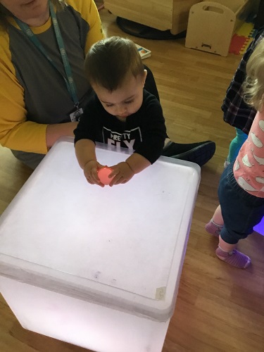 A young child standing with support from an educator next to a light cube