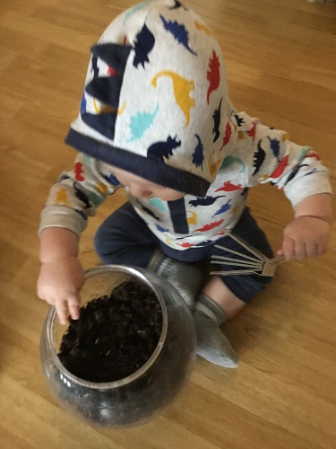 An infant sitting on the ground with a large clear container filled with soil in front of him