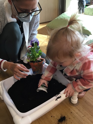 An infant scooping soil with a small cup next to an educator holding a flower in a pot