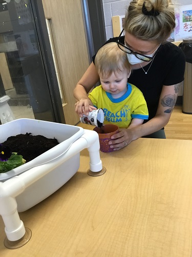 An educator and an infant scooping soil into a small pot at the table