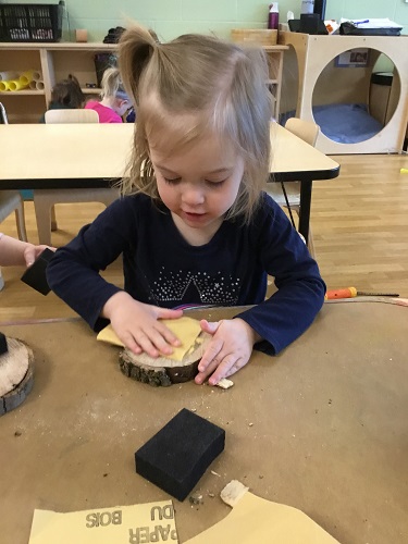 A child using sandpaper on a wood cookie in front of her at a table
