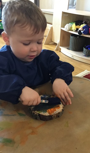 A child sitting at a table using a foam paint brush to paint the wood cookie in front of him