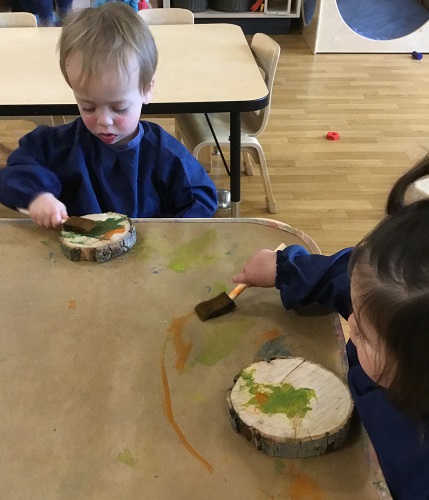 Two children sitting at a table using foam paint brushes to paint the wood cookies in front of them