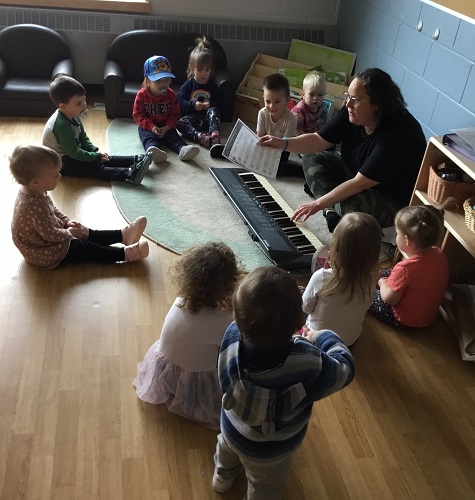 An educator and a group of children sitting on the floor listening to the educator play the keyboard