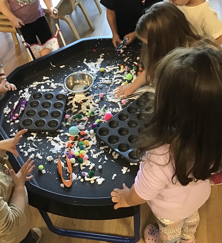 Children surrounding a tuff tray that has pompoms, tongs and cupcake trays on it