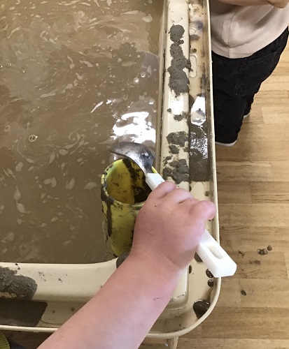 A child's hand holding a spoon scooping mud into a tube