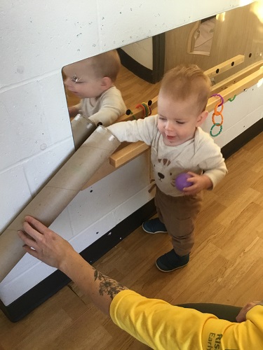 An infant reaching into a cardboard tube taped to the wall