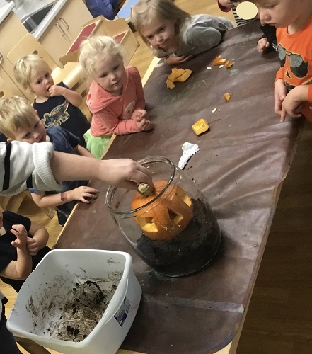 A group of children surrounding a table watching a pumkpin being placed in a large jar