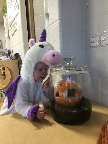 A child in a Halloween costume looking through a large jar at the pumpkin inside