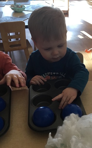 A child sitting at a table touching items in a muffin tin in front of him
