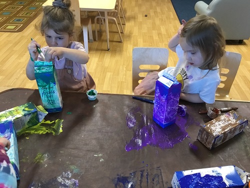 Two children painting milk cartoons at a table