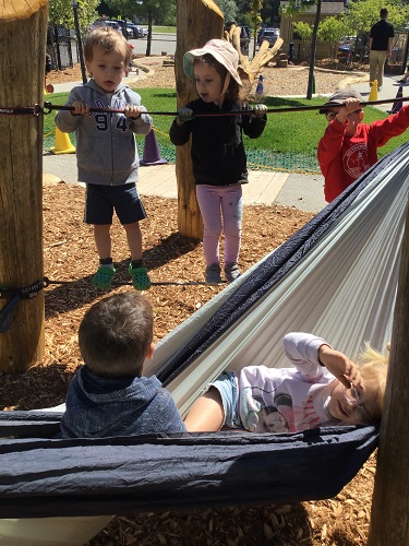 Two children standing on the ratchet straps attached to the wacky posts, looking at the children in the hammock