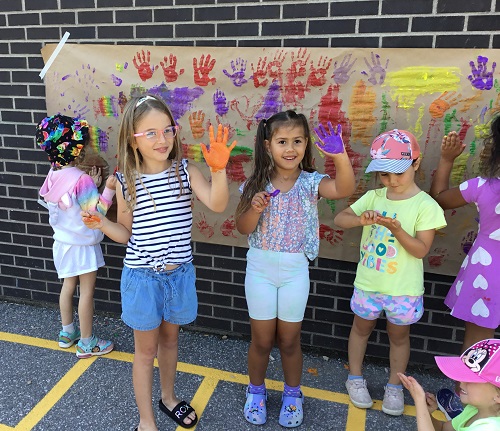 Children outside standing in front of a large paper stuck to the brick wall, showing off their painted hands. 