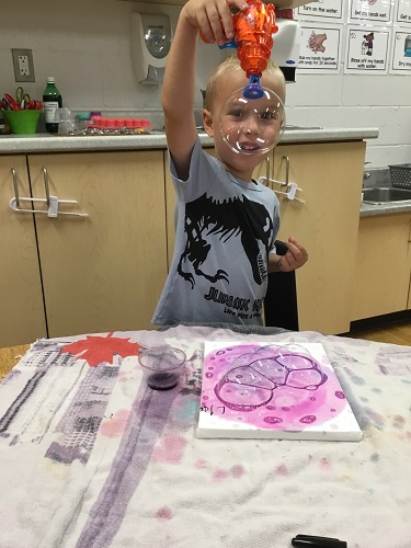 A child sitting at a table holding an upside down bubble gun with a large bubble in front of his face