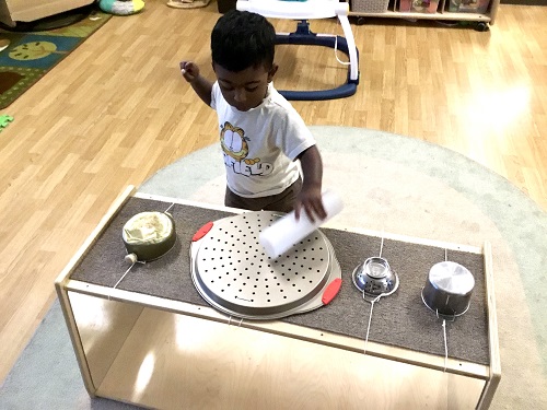 A child using a large tube to bang on a shelf covered in metal pots and pans