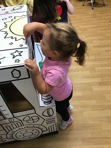 A child standing on her tip toes to colour the roof of a cardboard bus