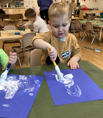 A child using a paint brush to spread fluffy white paint on a blue paper