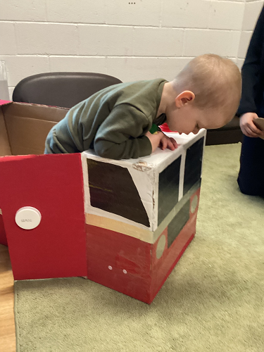 An infant peeking over the front of a fire truck made out of boxes