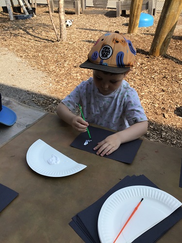 A child sitting at a table using a paint brush to spread sunscreen on a piece of black construction paper