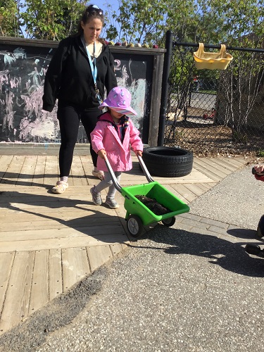 A child pushing a wheelbarrow filled with soil through the playground