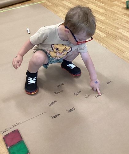 A child squatting down to point to his name written on the brown paper on the floor