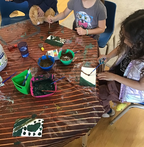 2 School age children sitting at the table with mud paint, brushing paint on their canvas with a paintbrush