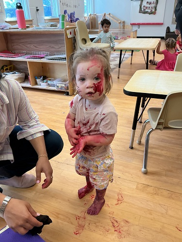 one Toddler standing head to toe covered in paint, there are 2 Toddler children in the background, one has their back turned, one is looking at the camera