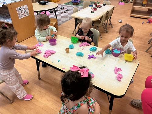 5 toddlers standing and sitting around the table with playdough and big cupcake holders manipulating the playdough