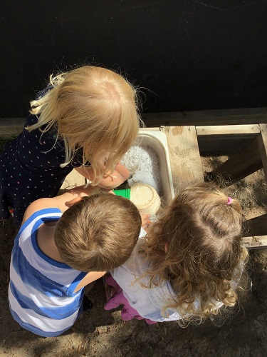 3 Preschool children standing around a small sensory bin with bubbly water using bowls to scoop the bubbles