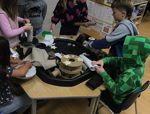 6 School Age 2 children standing and sitting around the table with a tuff tray filled with loose parts - constructing their creations