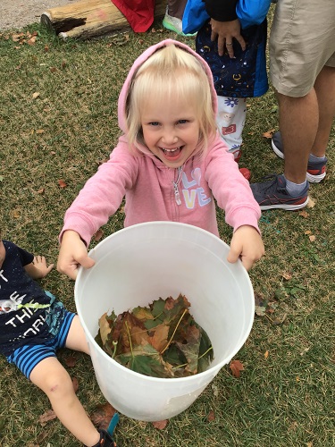 Preschool child holding a bucket filled with leaves and smiling big
